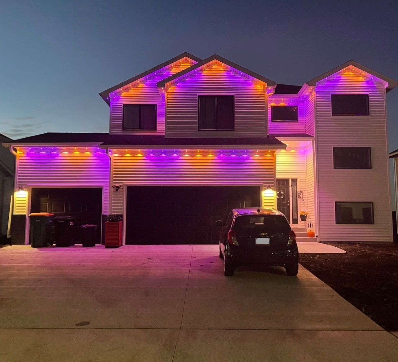 A suburban house at dusk decorated with purple and orange Halloween lights along the rooflines and a car in the driveway.