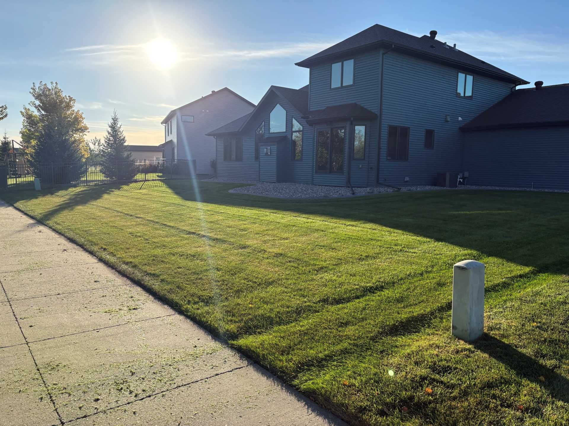 A sunny lawn in front of a blue two-story residential home with a sidewalk in the foreground.