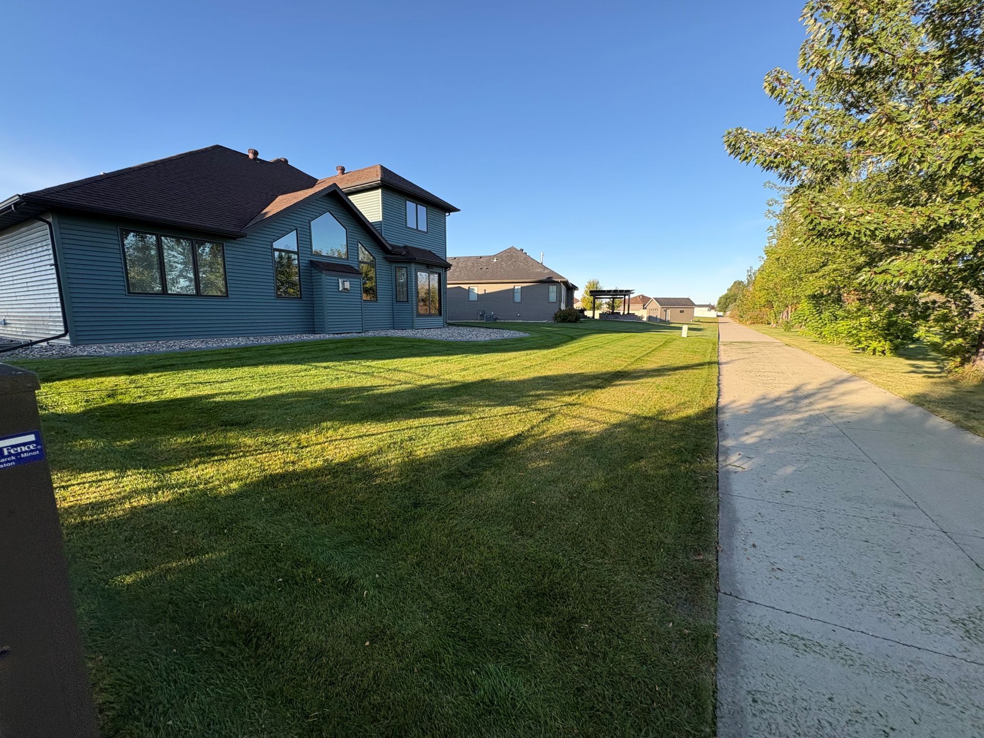 A multi-story dark gray house stands beside a paved walking path bordered by a grassy lawn and a row of trees.