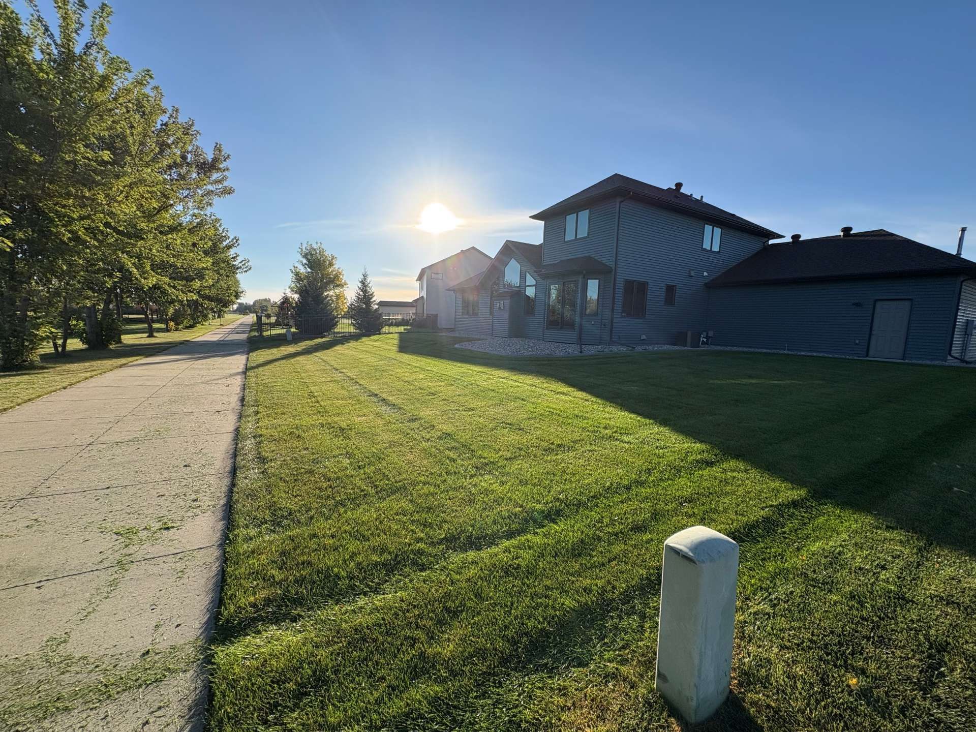 A sunny, clear sky overlooks a suburban house with dark siding, a grassy lawn, and an adjacent gravel path.