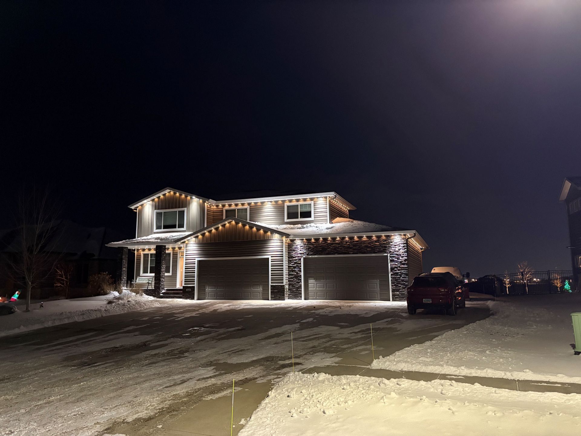 A two-story house at night with white festive lights along the roofline, set against a snow-covered yard and street.