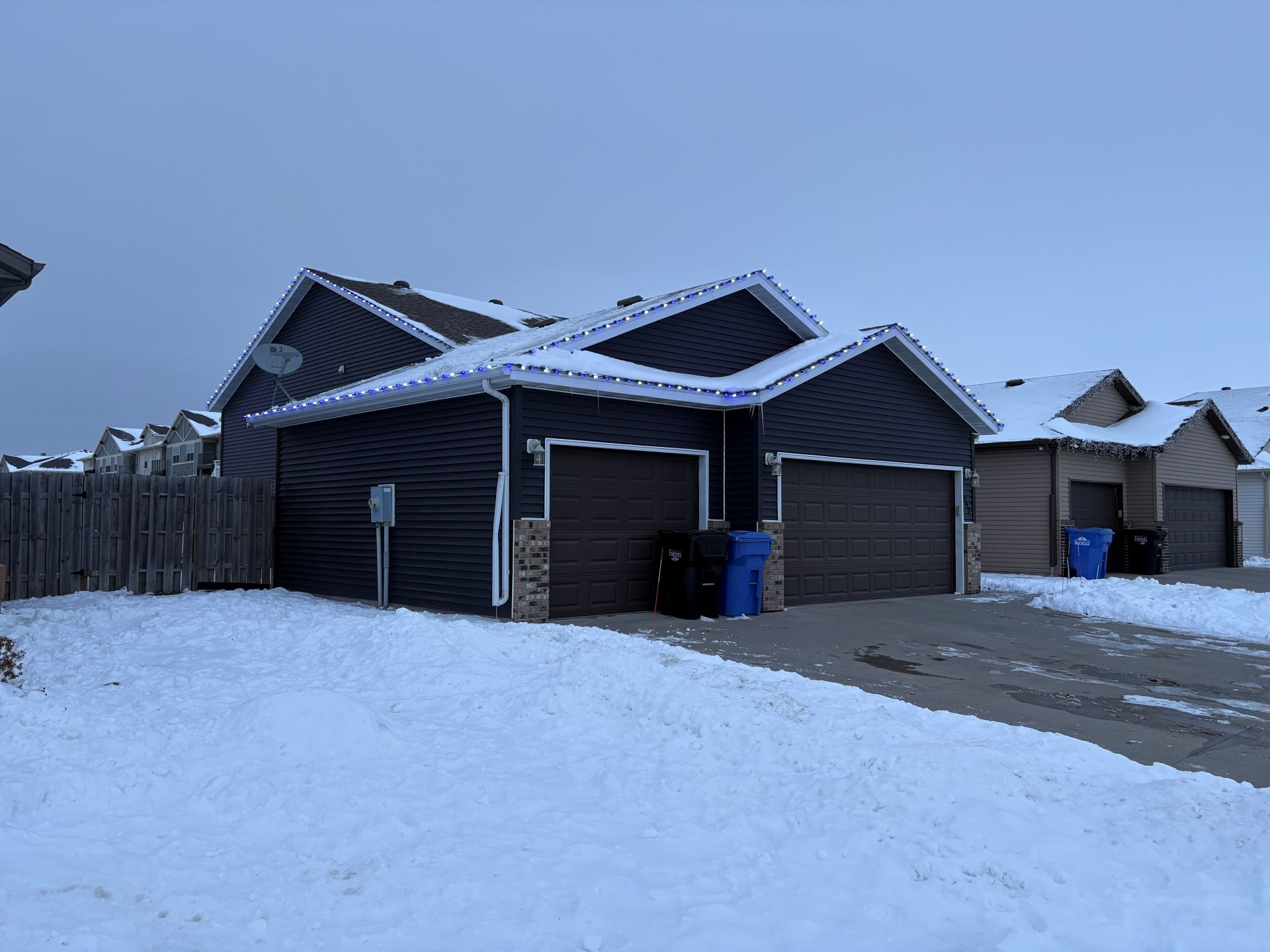 A dark blue, two-car garage duplex with snowy rooftops and driveways on a winter day.