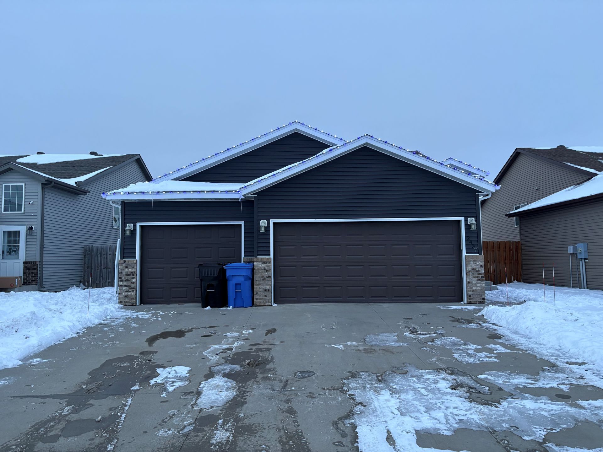 A suburban house with a dark blue exterior, two brown garage doors, and snow-covered ground and roof.