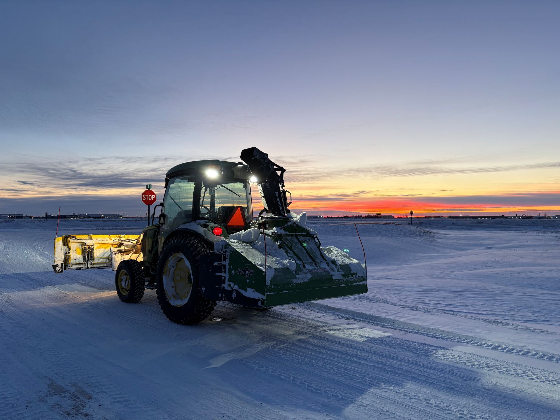 A snow removal tractor with a plow and snow blower operates on a snowy airfield during a colorful sunset.