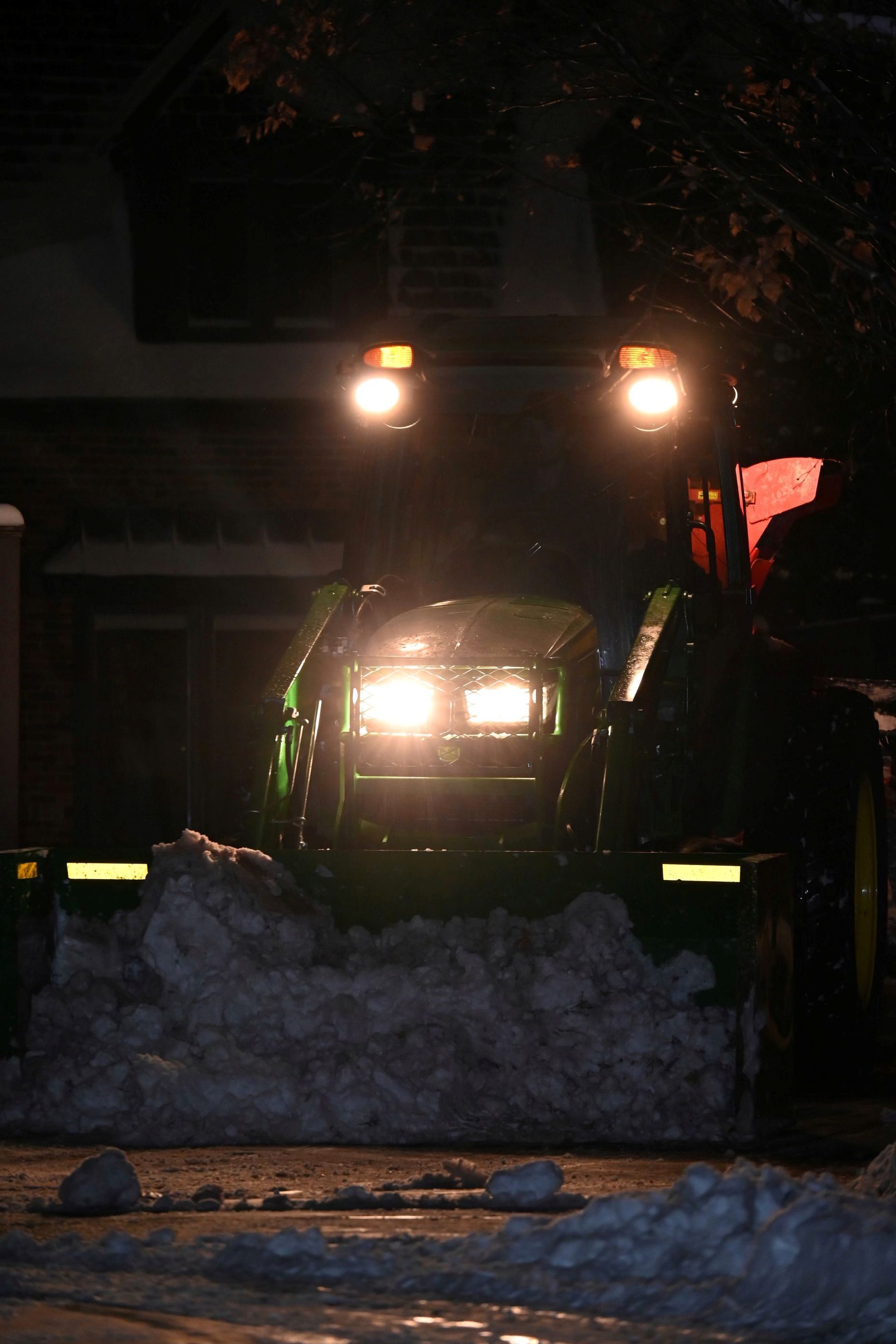 A green tractor with bright headlights clears a pile of snow on a road at night.