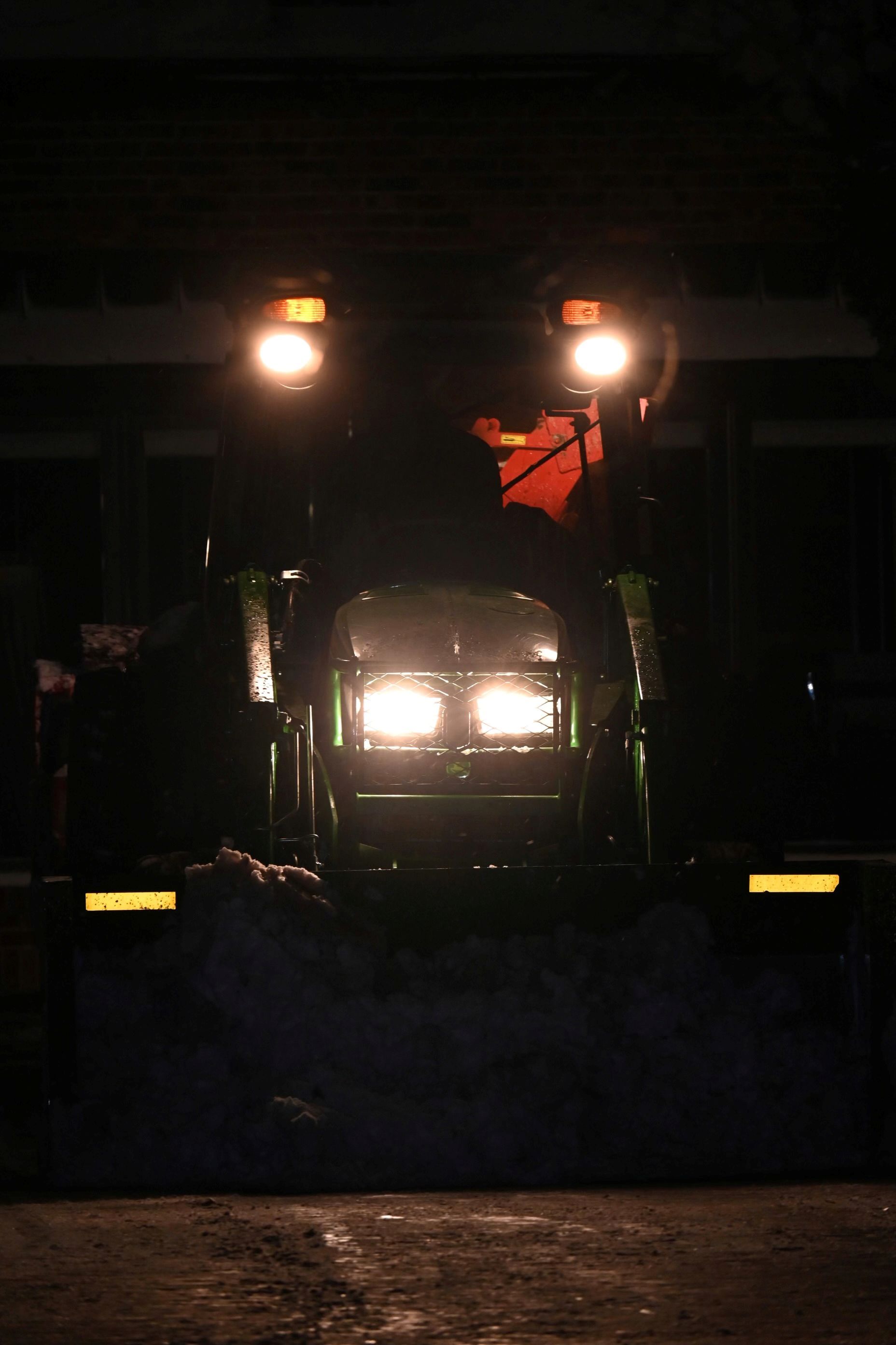 A tractor with bright headlights illuminates a dark, snowy setting at night.