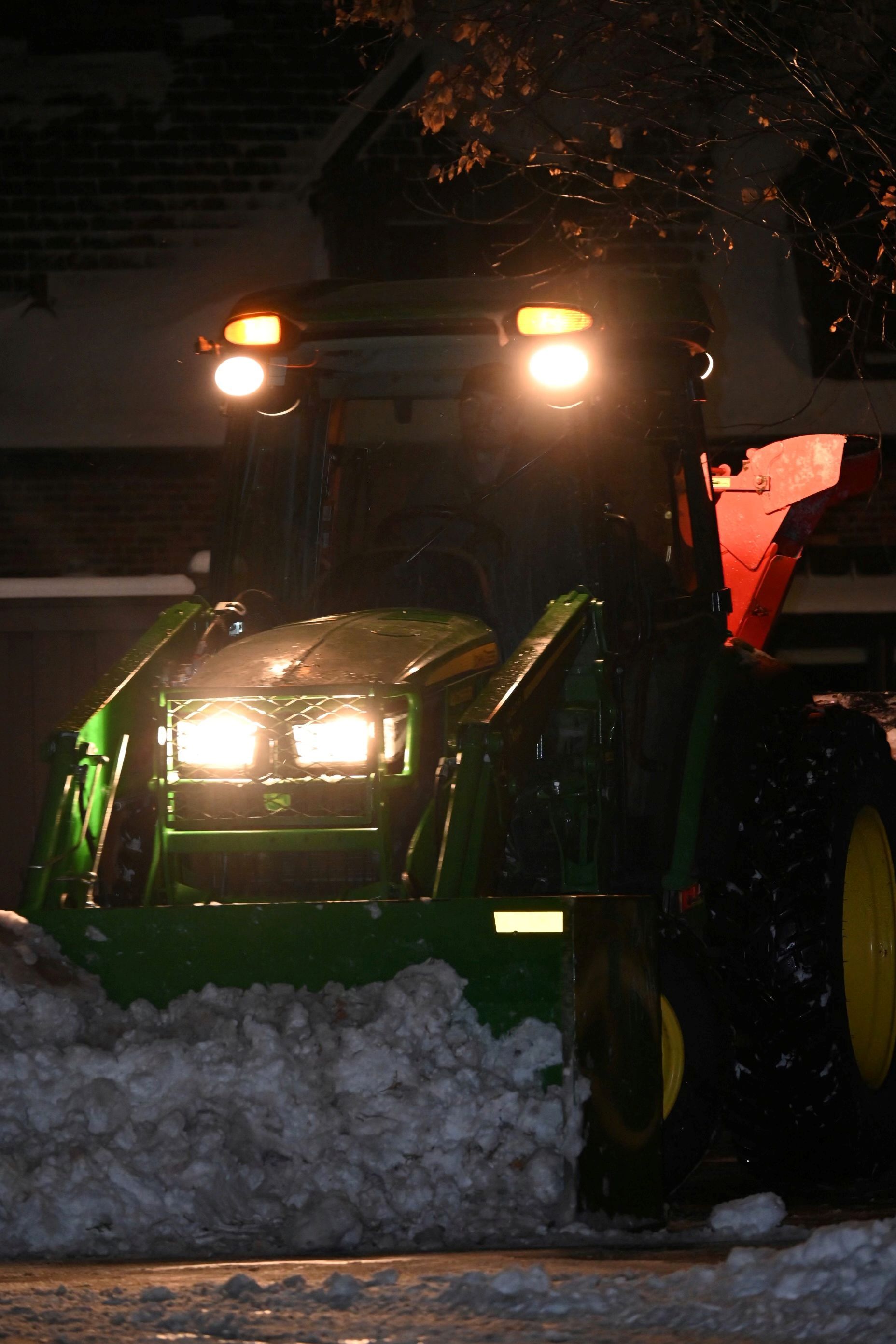 A green John Deere tractor with bright headlights clears snow at night.