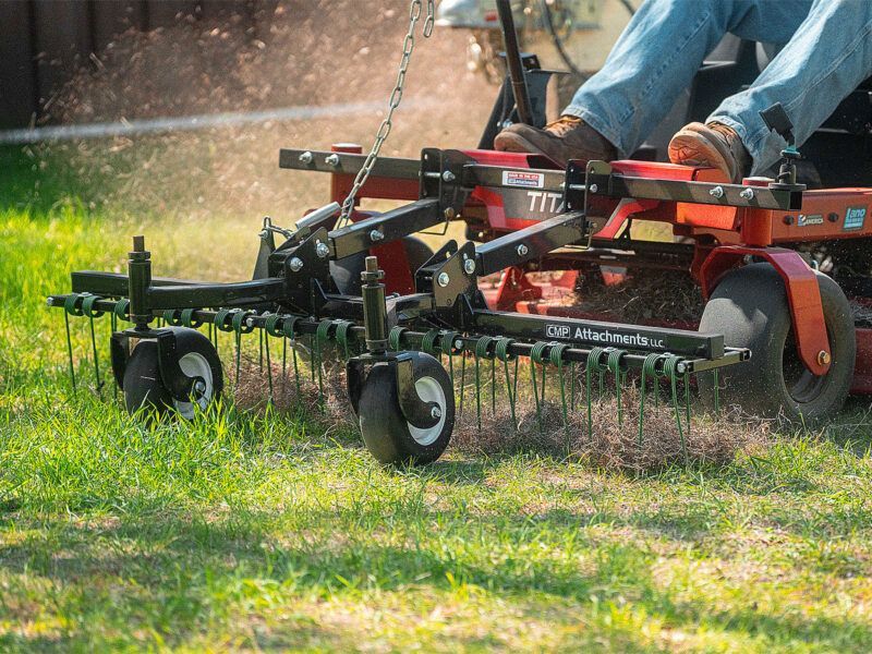A mower attachment with small wheels and metal tines detaches dead grass from a lawn, pulled by a red riding mower.