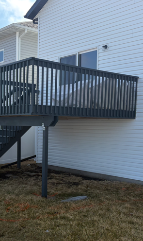 A low-angle view of a grey wooden deck attached to the back of a two-story white house with light-colored siding.