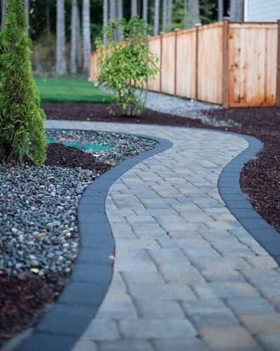 A curved walkway made of grey pavers with a dark border, bordered by decorative stones and mulch in a landscaped yard.