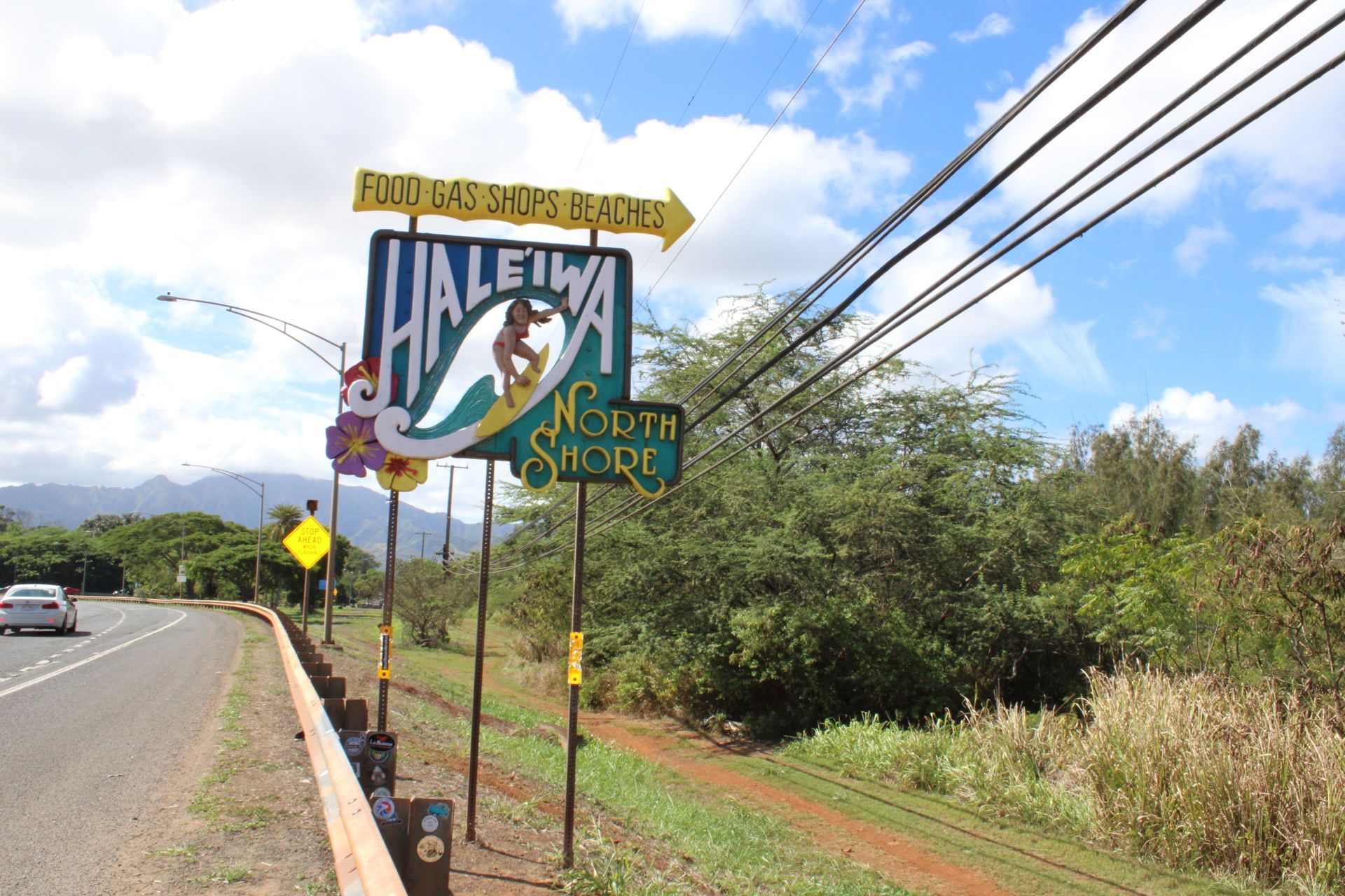 Haleiwa North Shore sign with a surfer graphic. Roadside view with car and trees under cloudy sky.