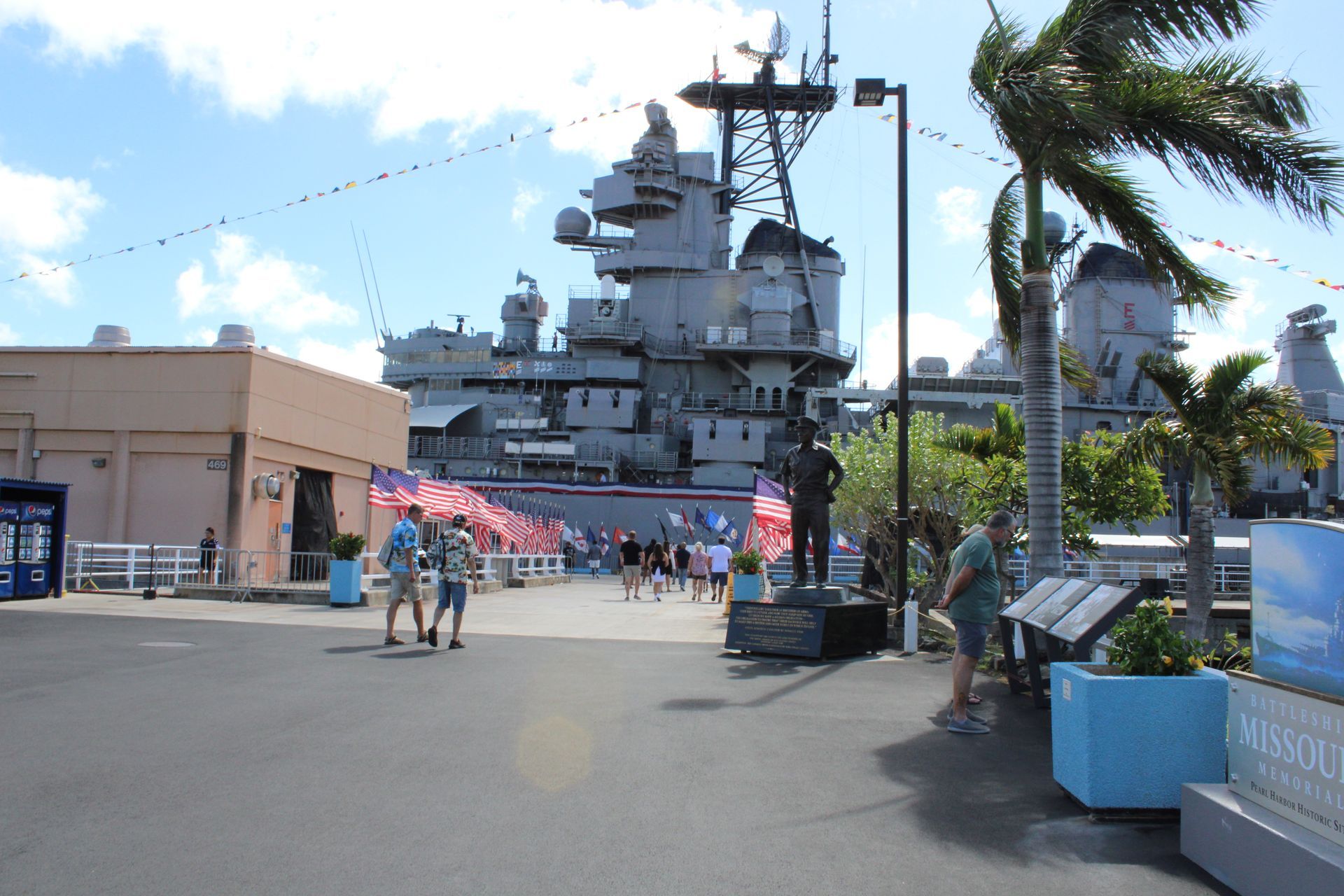 People walking towards a large battleship in a sunny outdoor setting with palm trees and a statue.