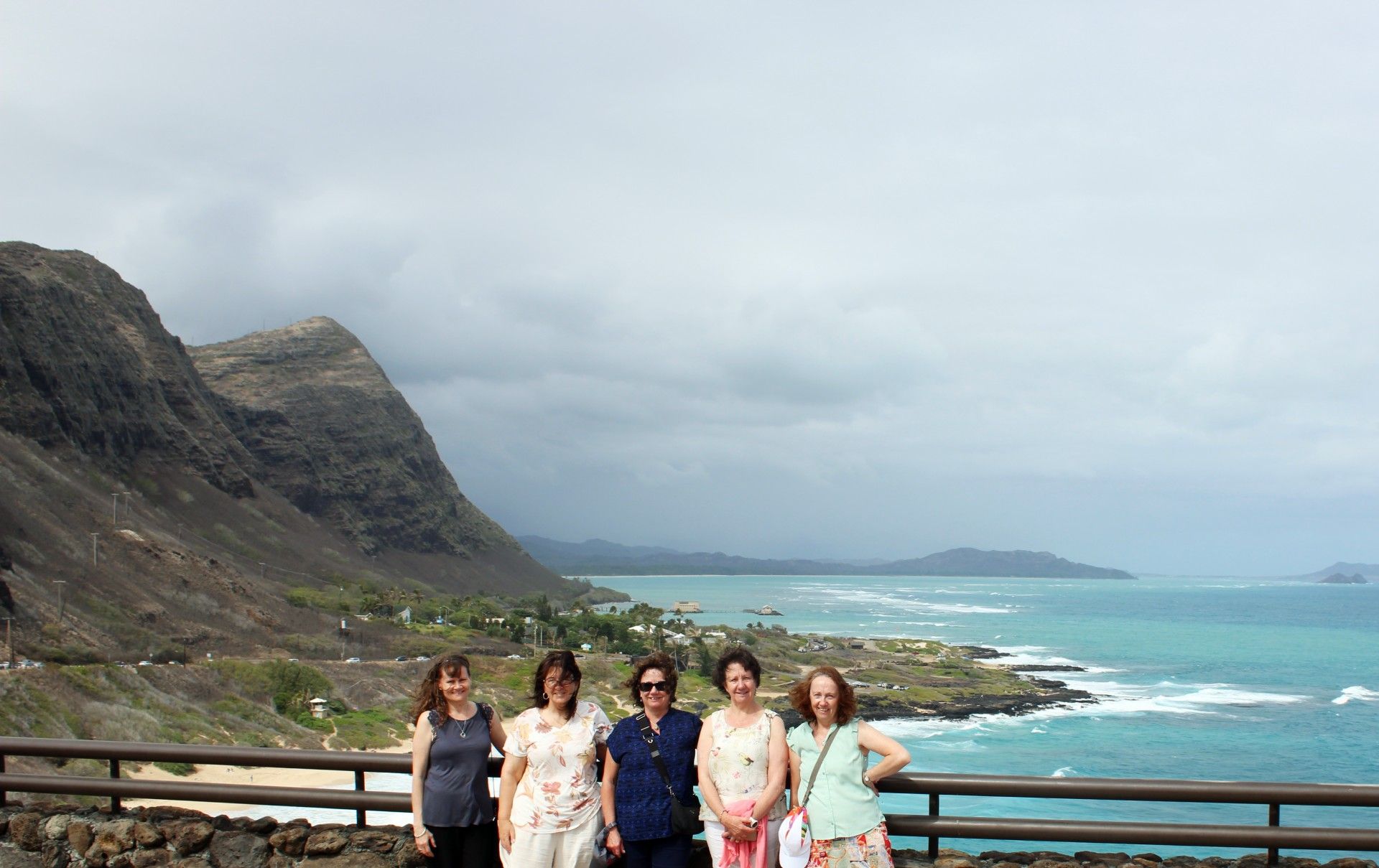 Five people posing by a scenic coastline with mountains, overcast sky, and turquoise water.