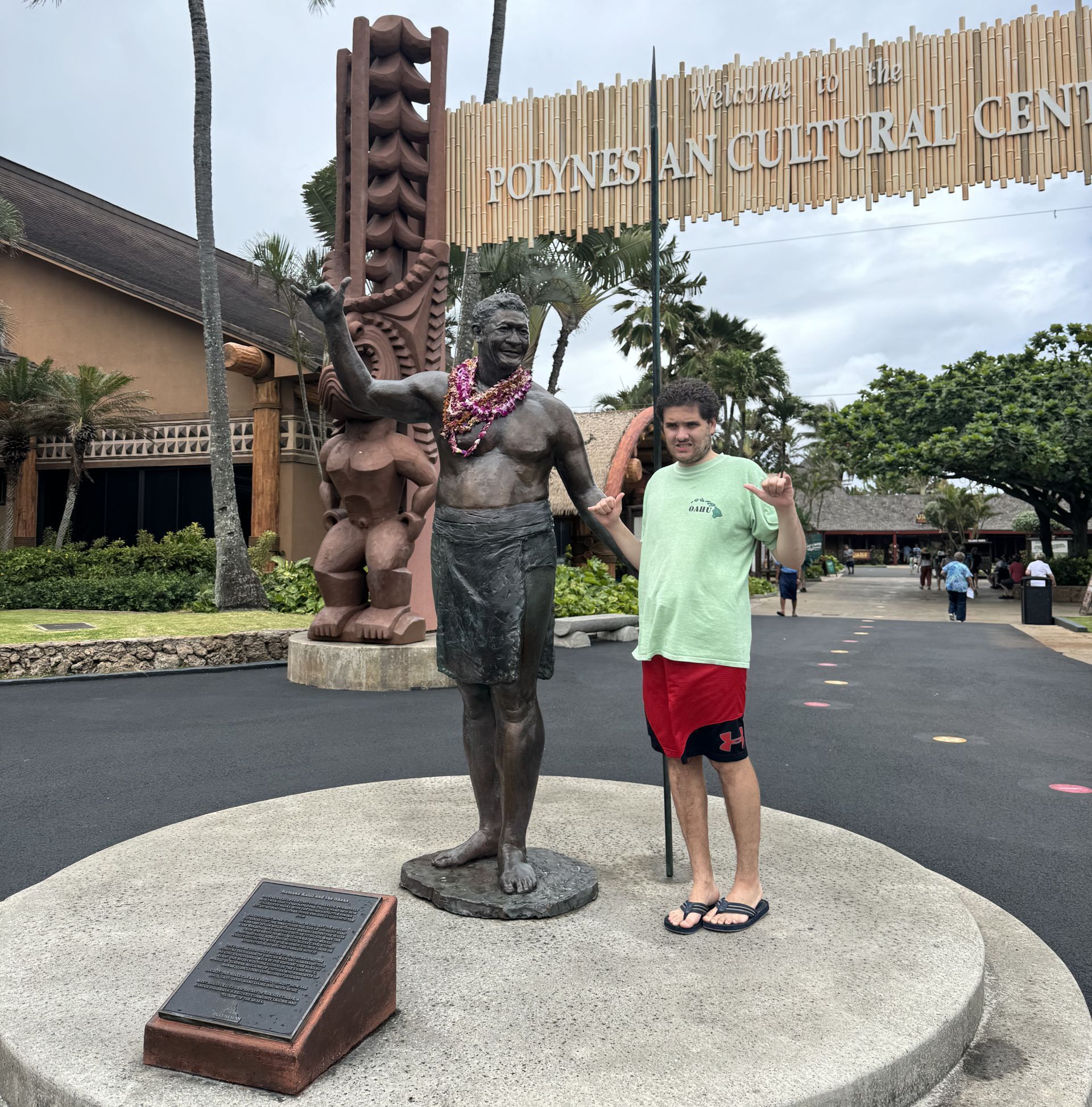 Man posing with a statue at Polynesian Cultural Center in Hawaii. Wearing red shorts and green shirt, arms raised.