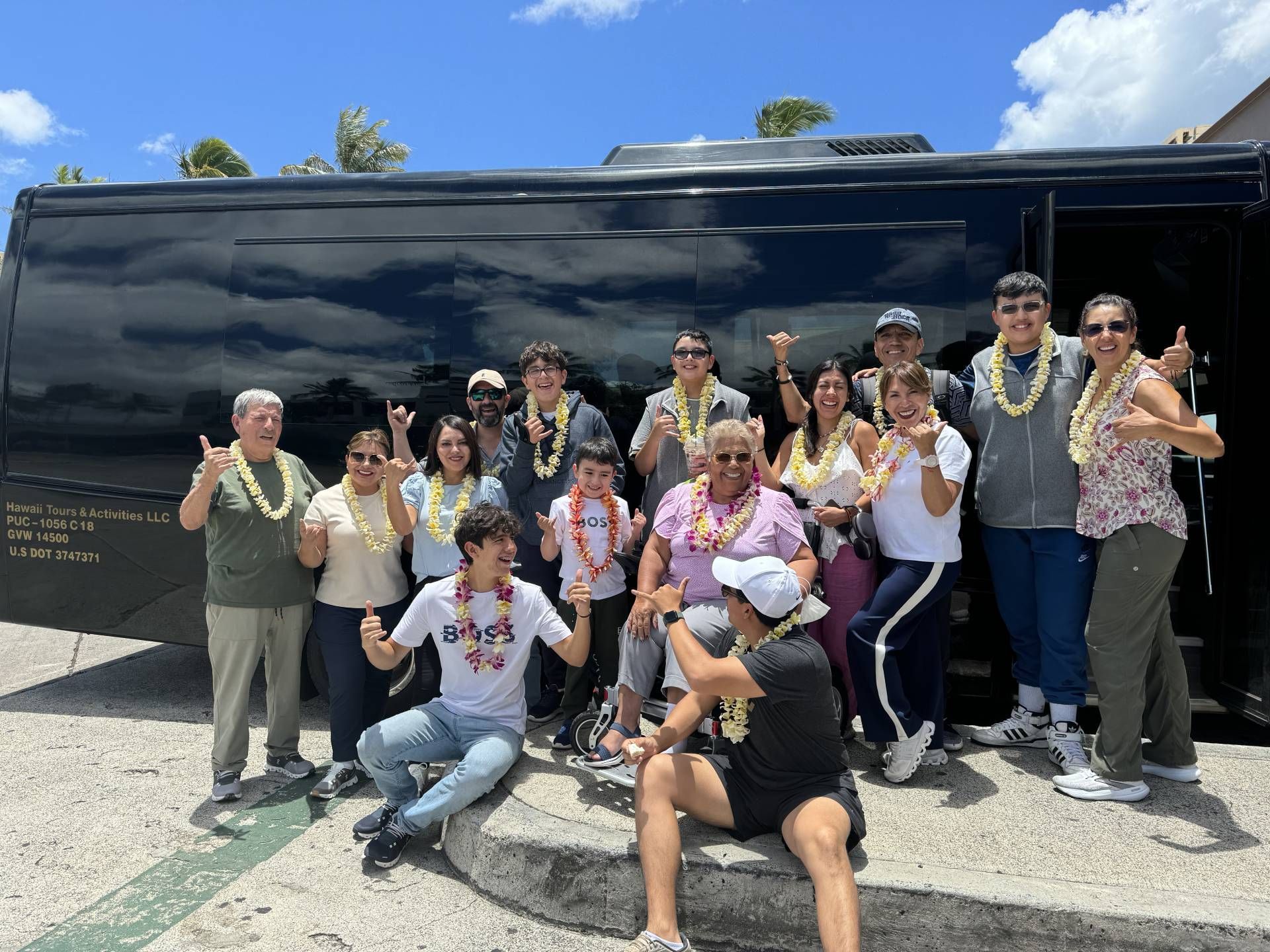 Group of people wearing leis poses in front of a black bus on a sunny day.