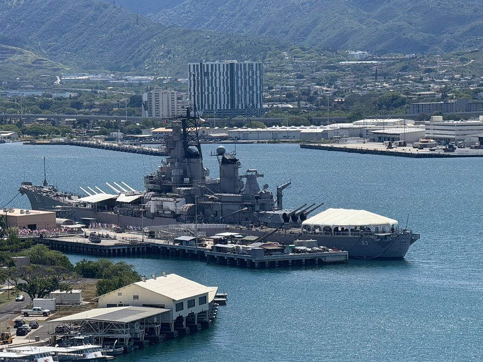 Battleship in harbor, docked at a pier. Buildings and a mountain are in the background.