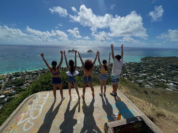 Man posing with a statue at Polynesian Cultural Center in Hawaii. Wearing red shorts and green shirt, arms raised.