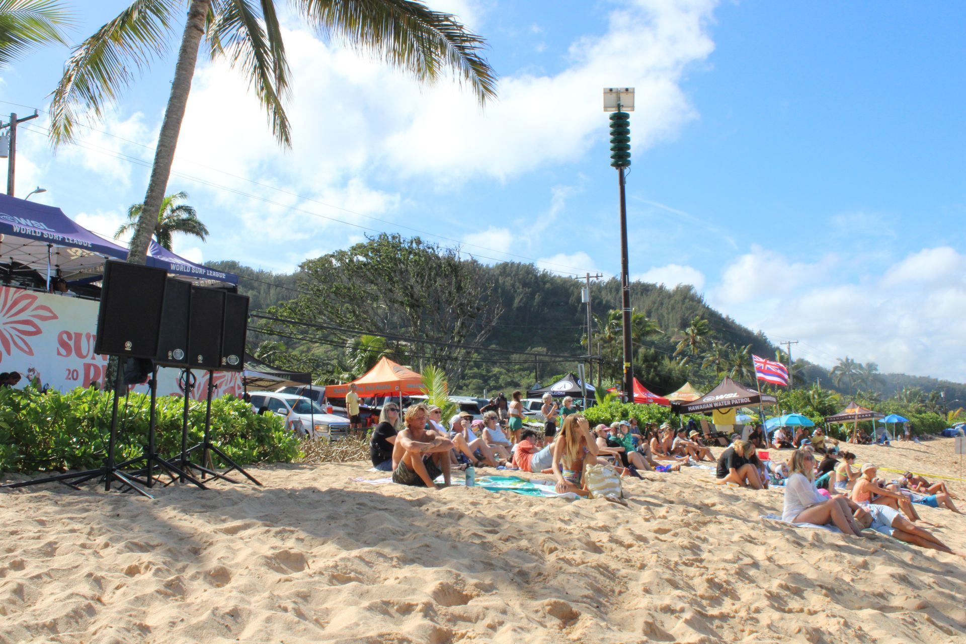 People sit in the sand, watching a surfing competition