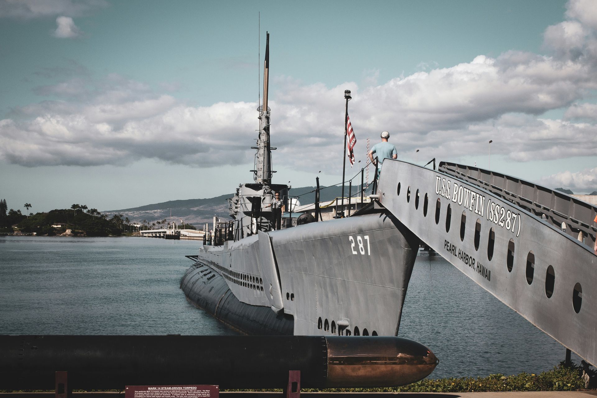 Submarine docked at a pier, numbered 267. Ramp leads from vessel, waterfront, blue sky with clouds.
