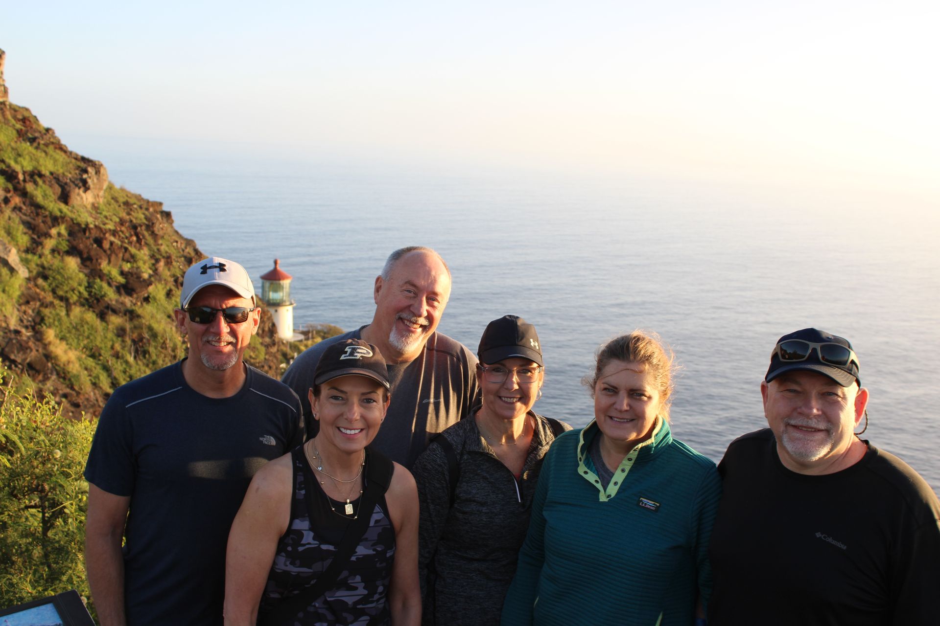 Group of people wearing leis, posing by a black bus; many are making shaka signs and smiling.