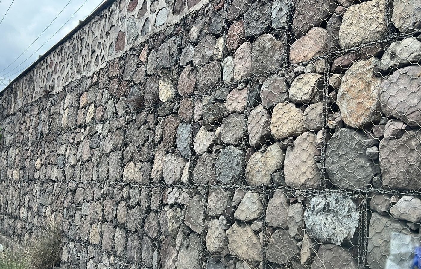 Muro de piedra con malla metálica, al aire libre, cielo nublado.