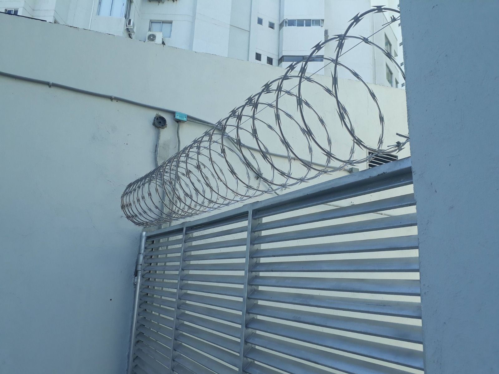 Bobinas de alambre de púas sobre una puerta y un muro metálicos, presumiblemente por motivos de seguridad en un entorno urbano.