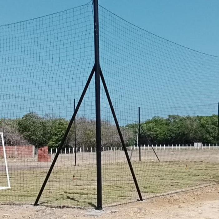 Una valla de malla negra sostenida por postes y puntales negros se alza sobre un campo de hierba, bajo un cielo azul.