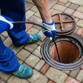 Plumber in blue overalls and gloves using a snake tool on an open sewer manhole.