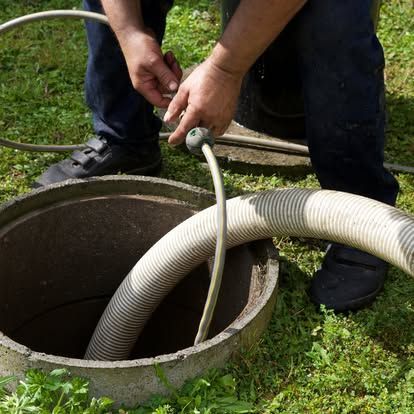 Person in dark pants and shoes holding a hose over an open concrete sewer cover in a grassy yard.