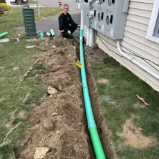 Man kneeling by a trench with a green pipe, likely for plumbing work, near a house's electrical box.