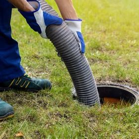 Person in gloves holding hose over open septic tank on a grassy lawn.