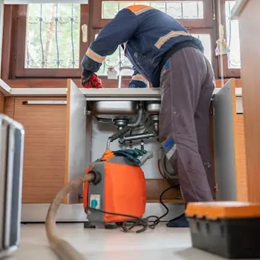 Plumber working on sink in kitchen, using pump. Orange and blue work clothes, gray pants.