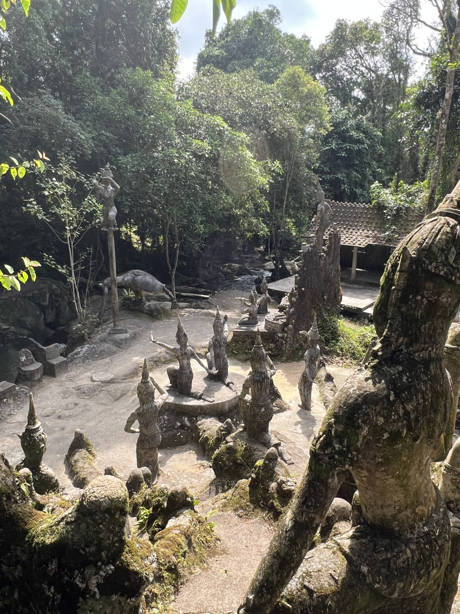 Stone sculptures in a lush garden setting; figures of various sizes stand on a sandy area, surrounded by trees.