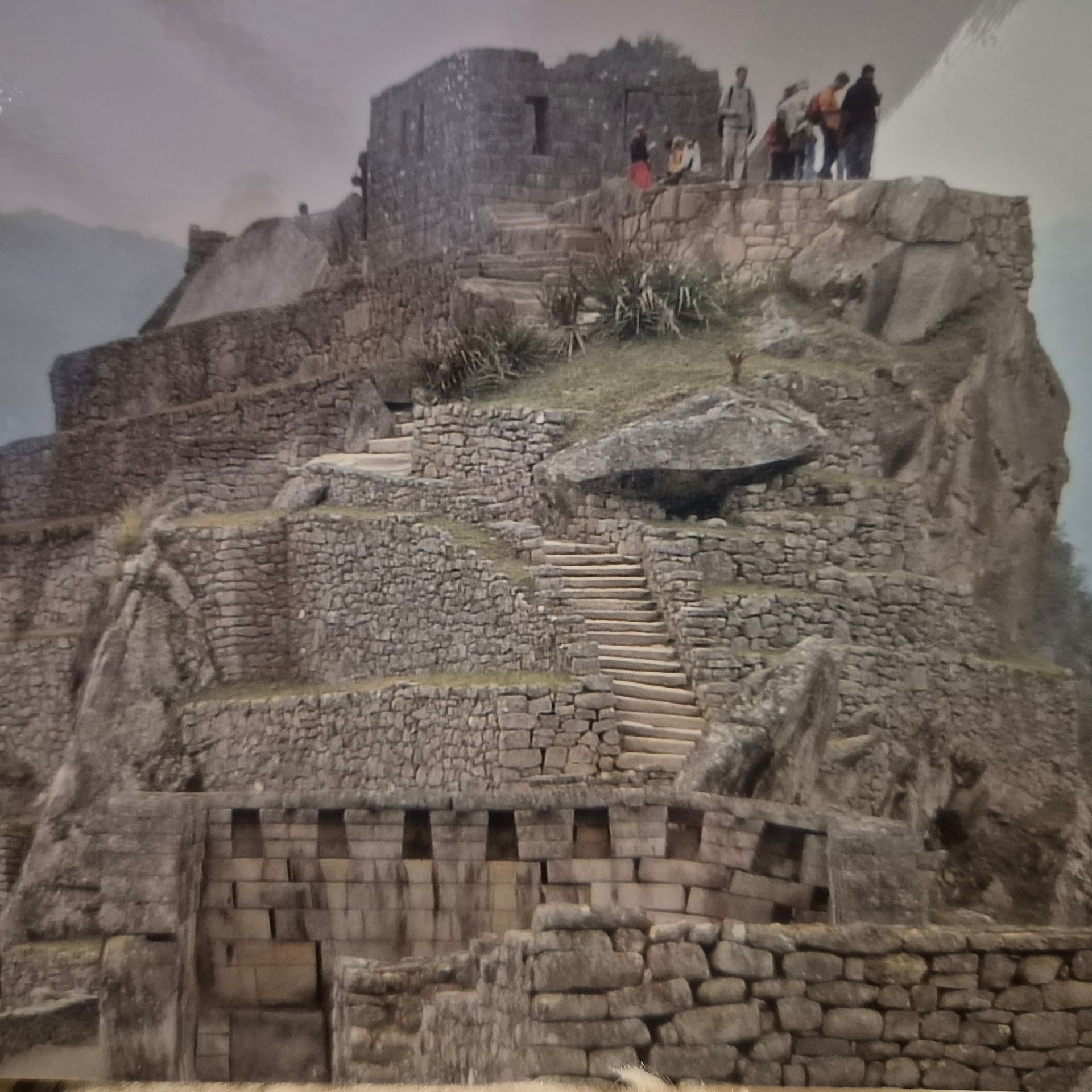 Machu Picchu ruins on a mountainside, with stone structures and stairs. People stand on the top looking at the view.