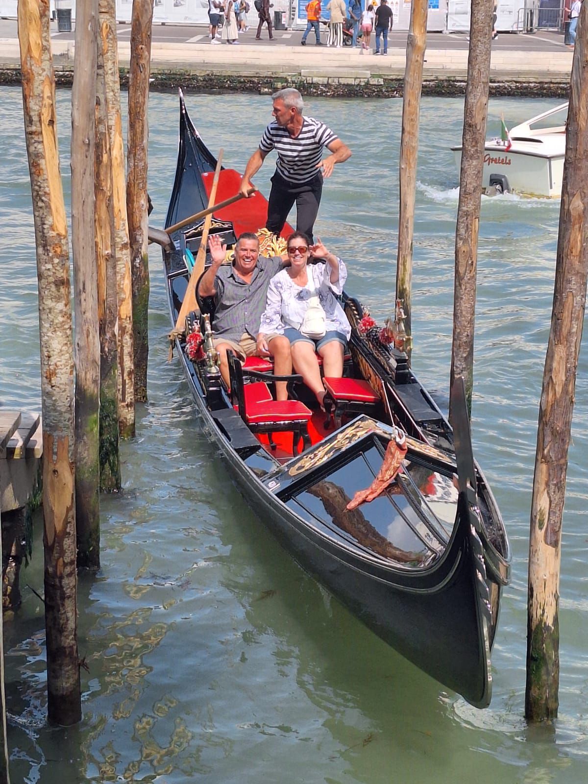 Gondola with three people in Venice: two passengers waving, a gondolier standing at the back, wooden posts on either side.
