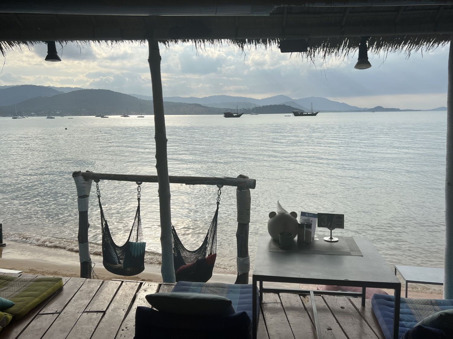 View from a beach hut: two hammock chairs, a table, and a seascape with boats and mountains. Cloudy sky.