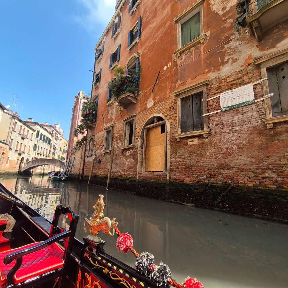 A gondola travels through a canal in Venice, passing aged buildings with red brick and wooden shutters.