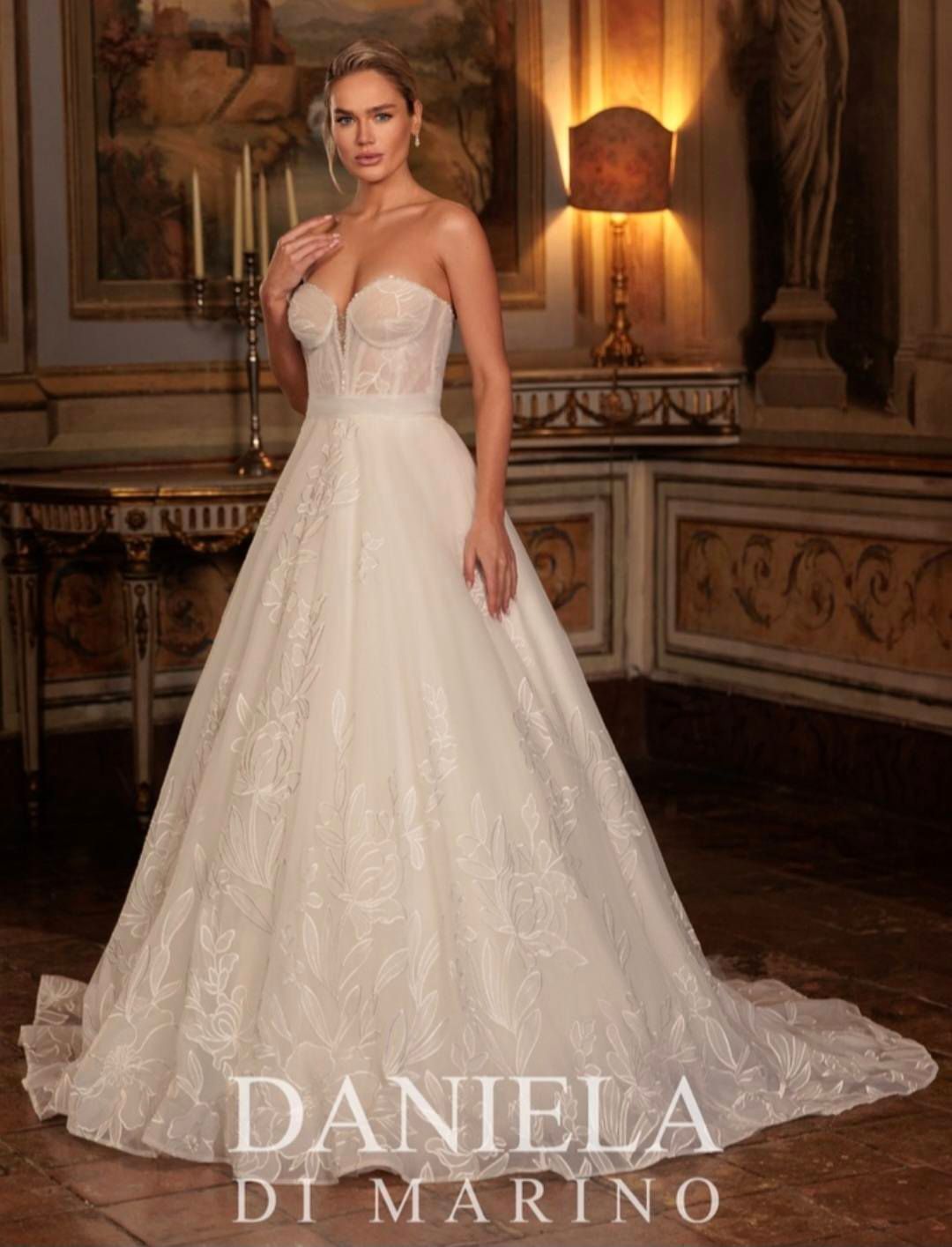Woman in a white wedding dress with long, lacy sleeves stands on a wooden dock near a lake.