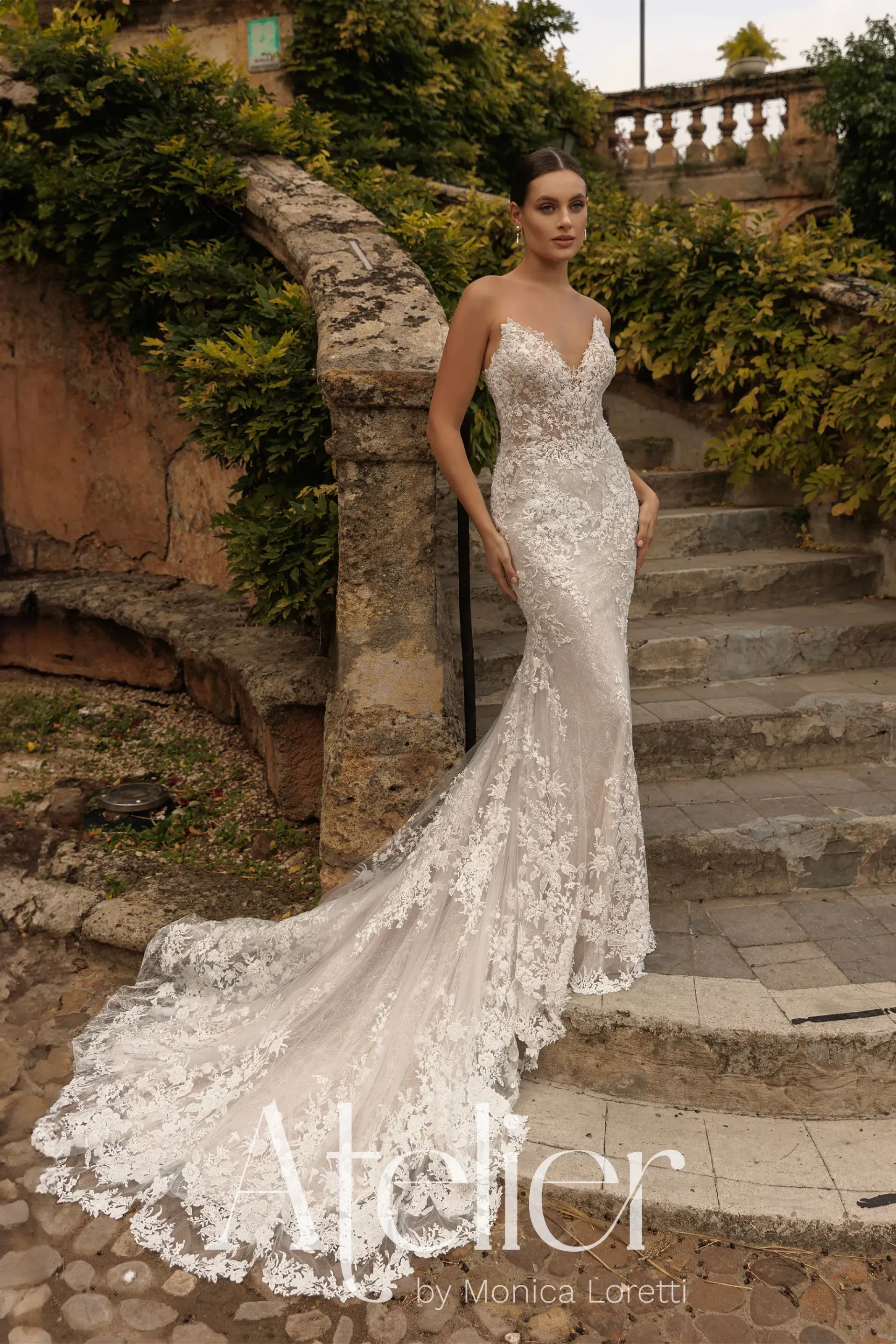 Woman in a white lace wedding dress with a long train, standing on stone steps outdoors.