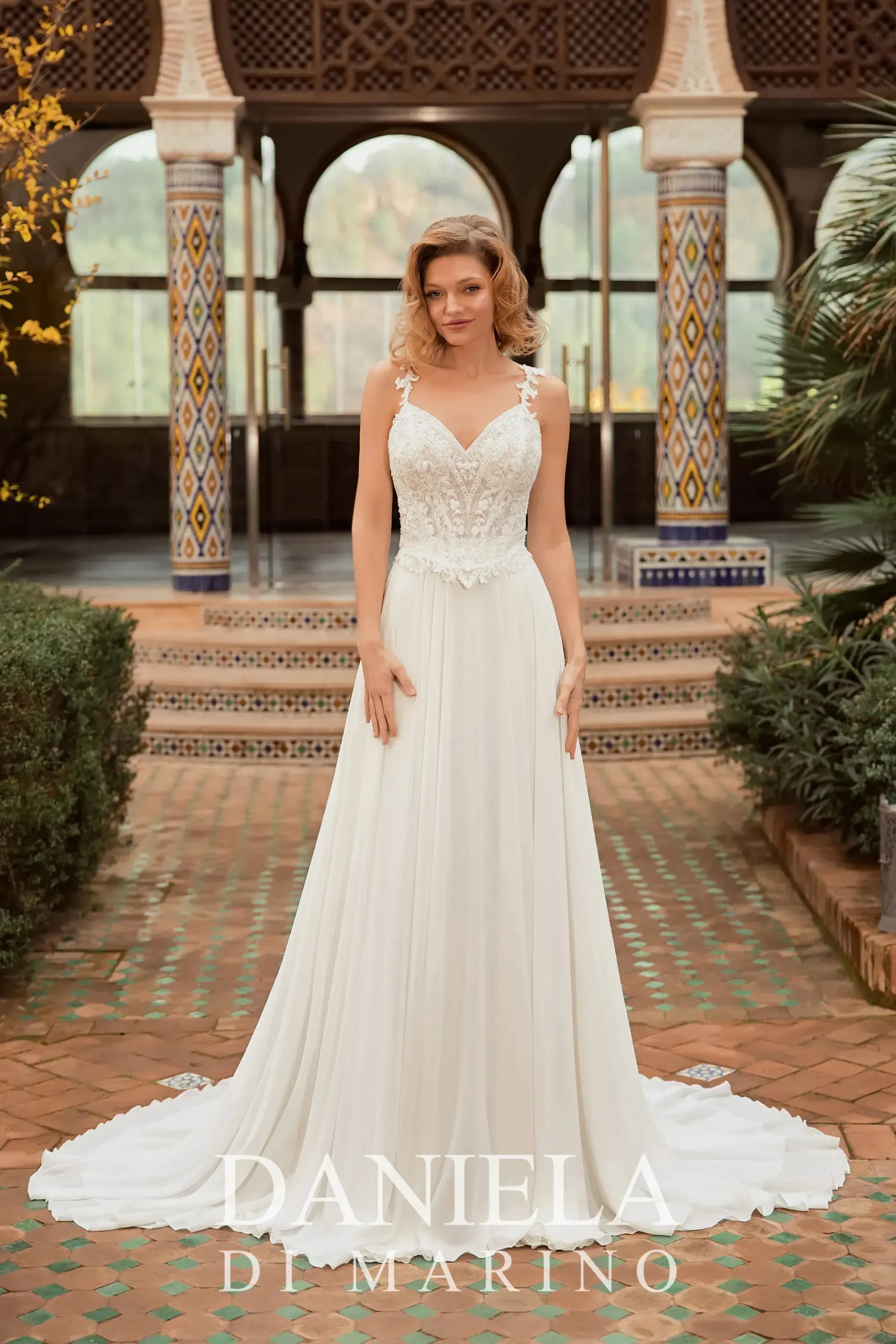 Blonde woman in a white wedding dress stands outdoors near pillars, with a floral bodice and flowing skirt.