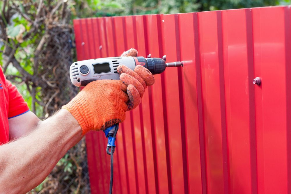 Person in Orange Gloves Using a Drill to Attach a Screw to a Red Metal Fence — Dachboe Your Handyman In Townsville, QLD