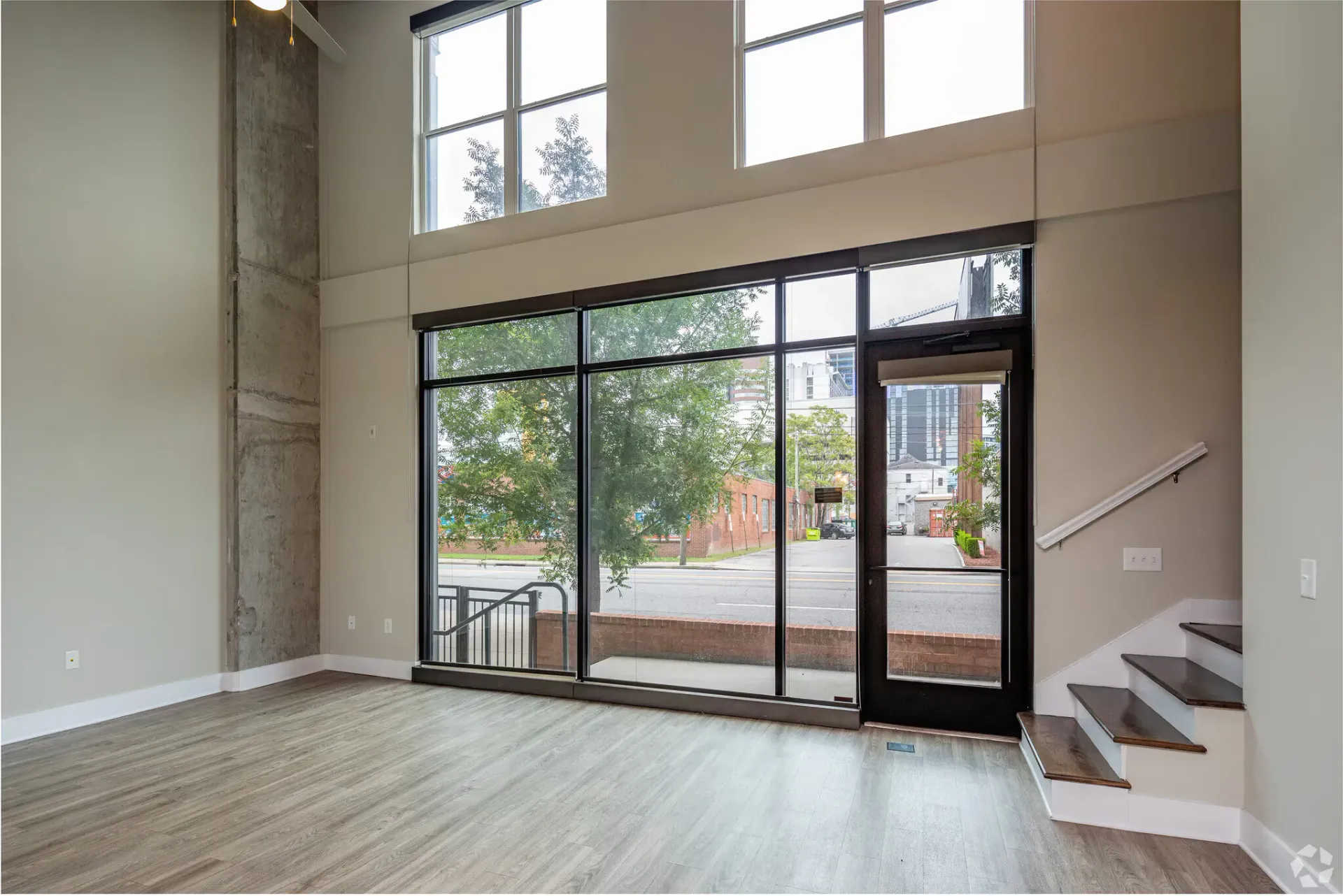 Bright living area with tall windows, glass door, concrete column, and staircase at the entry.