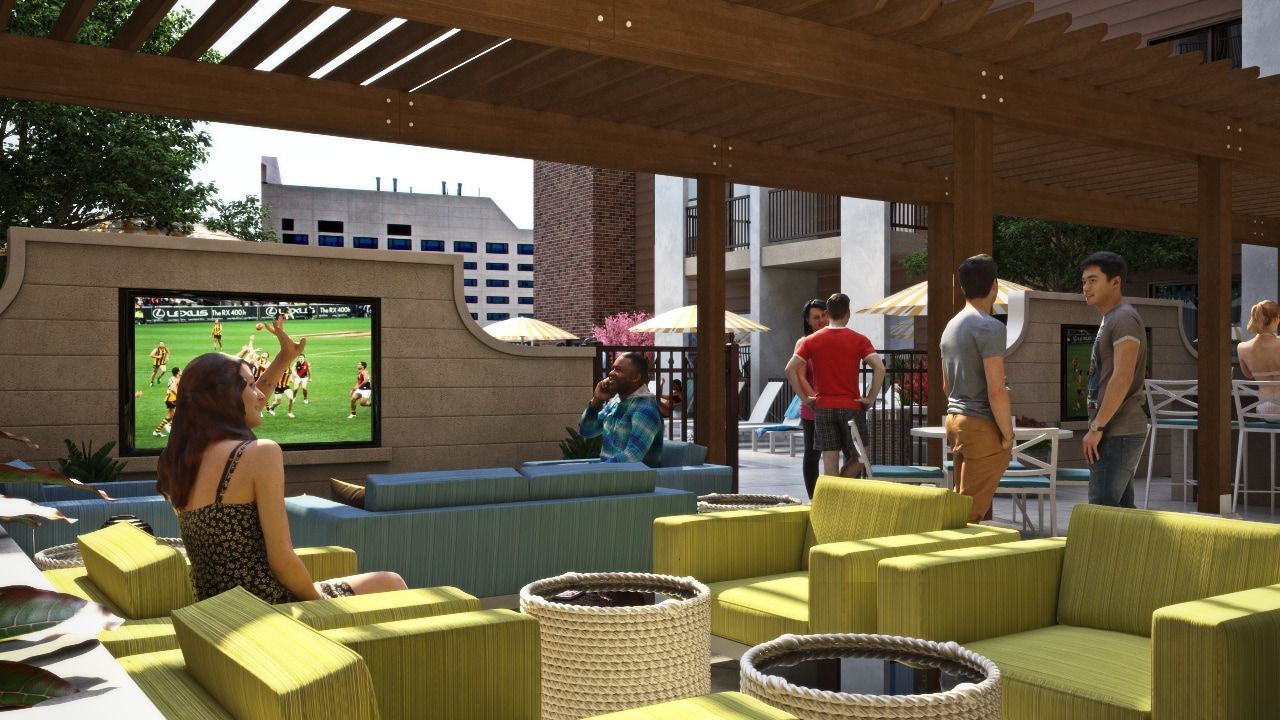 Outdoor communal lounge under a wooden pergola with colorful seating and a large wall TV, with people nearby.