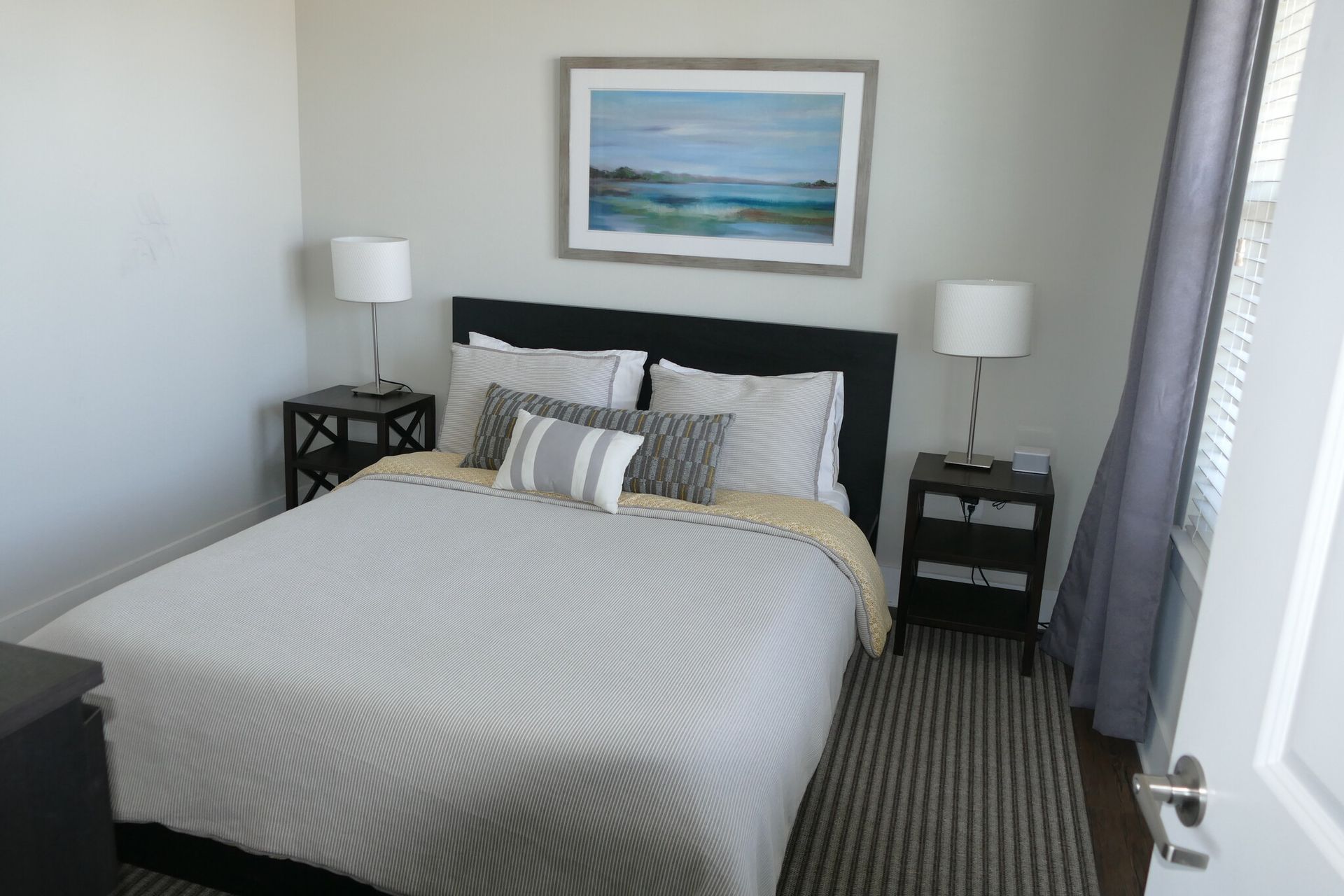 Bedroom in an apartment with a queen bed, two nightstands, lamps, and a framed landscape painting.