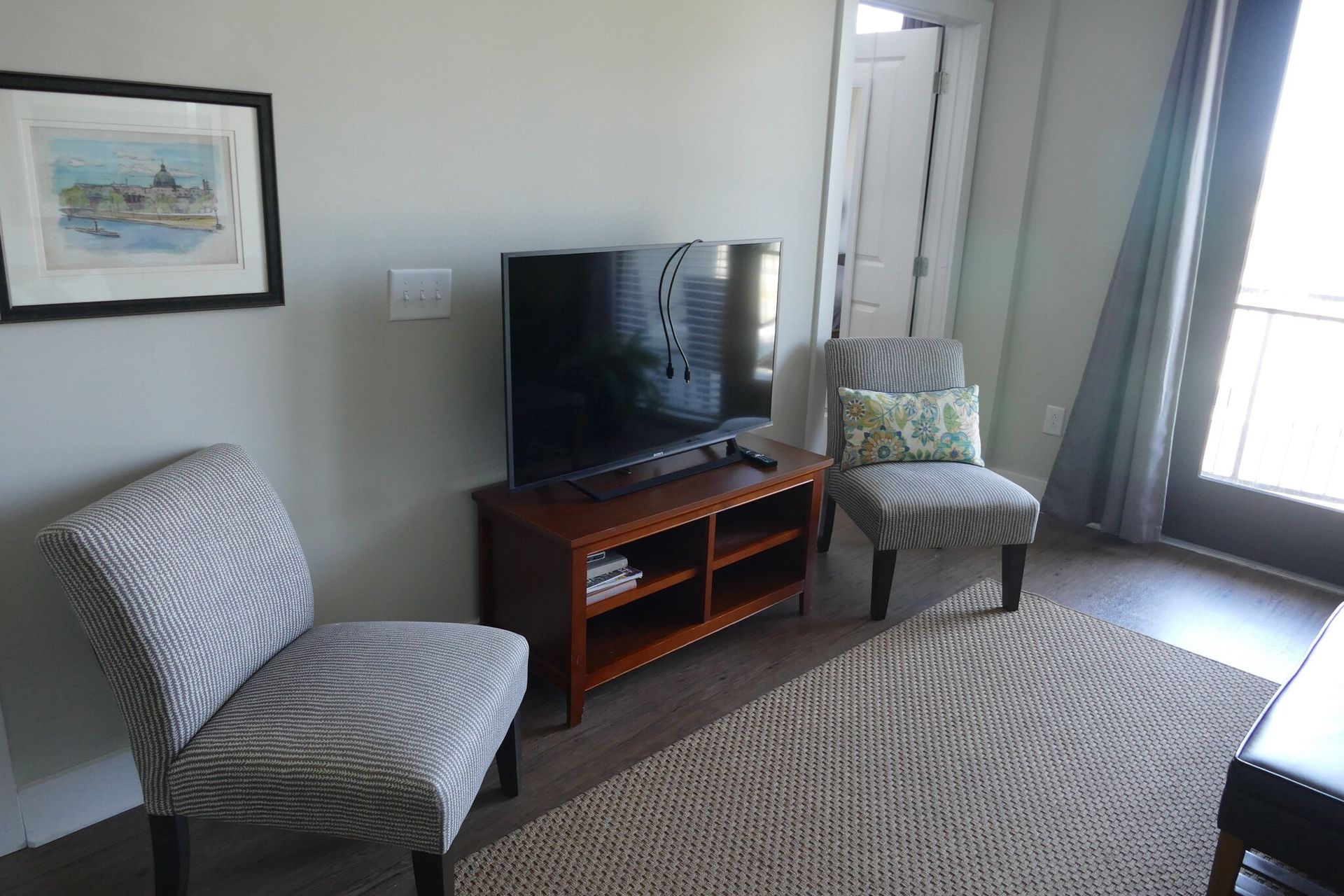 Living room with two striped chairs, a TV on a wooden stand, framed art, and a balcony door.