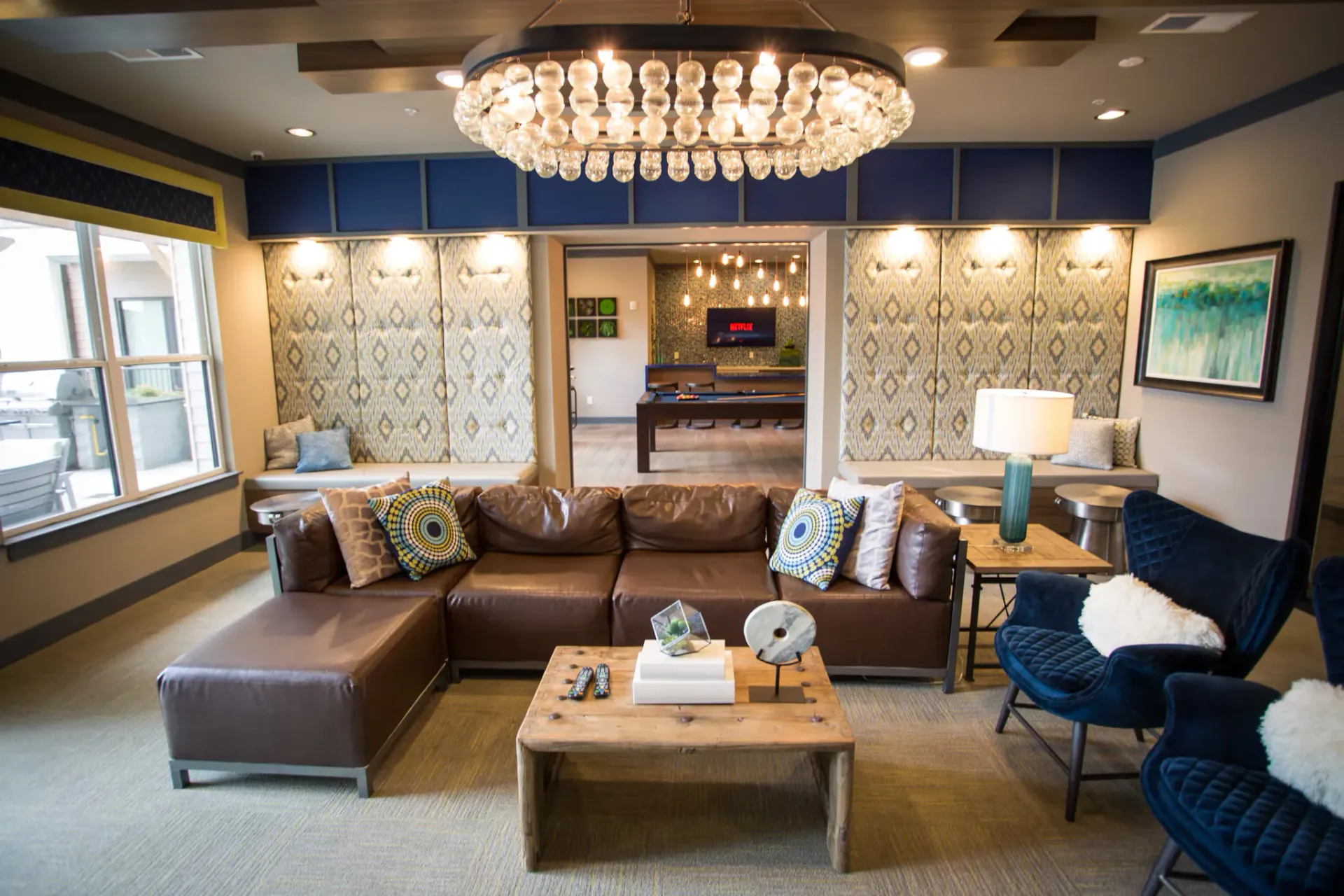 Communal lounge with a brown leather sectional, blue chairs, and a game area in the back.