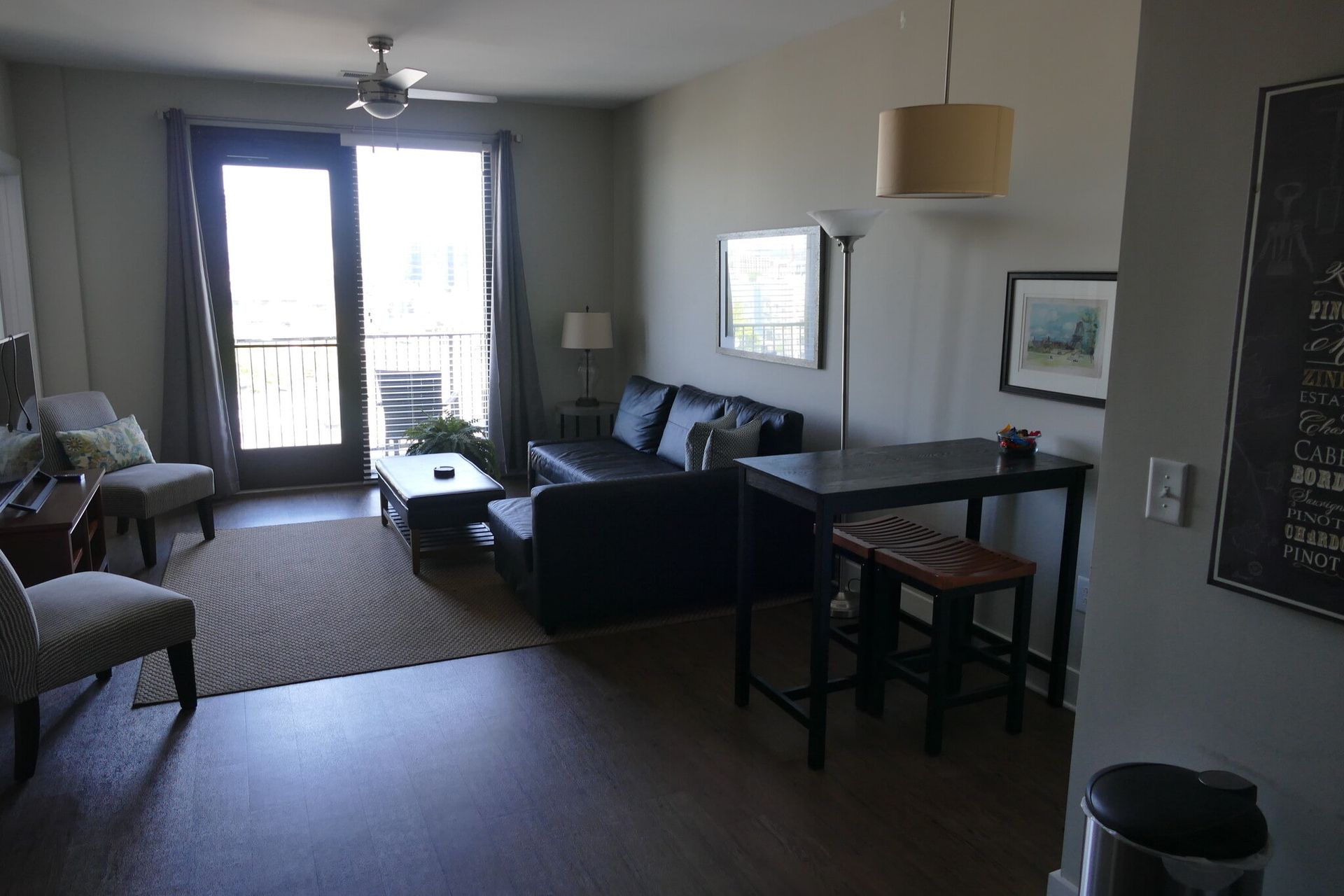 Living room in an apartment with a dark sofa, coffee table, balcony door, and a high dining table.