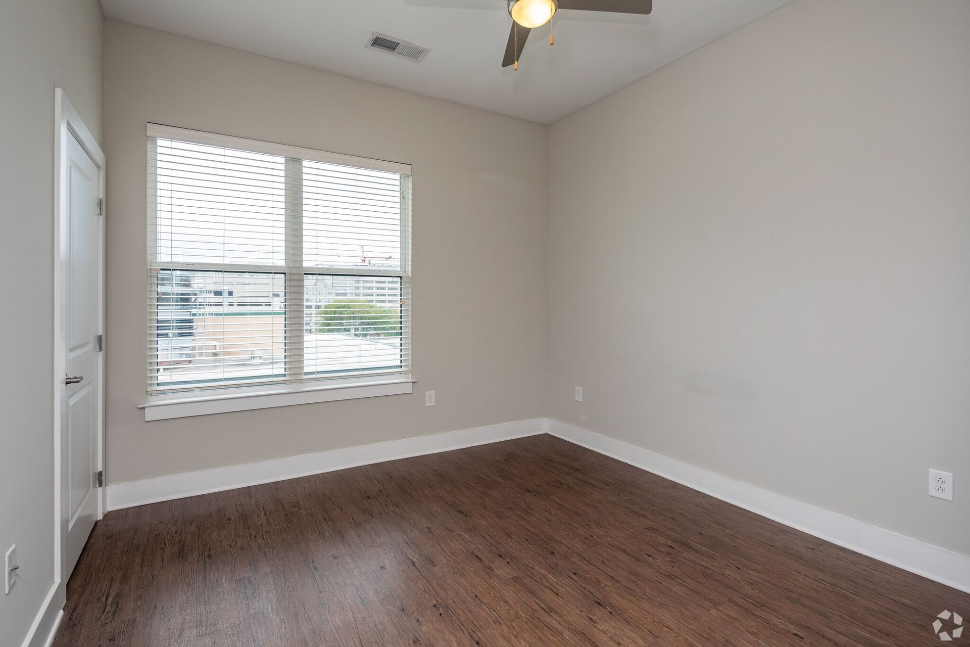 Empty bedroom with beige walls, dark wood-look flooring, large window with blinds, and a ceiling fan.