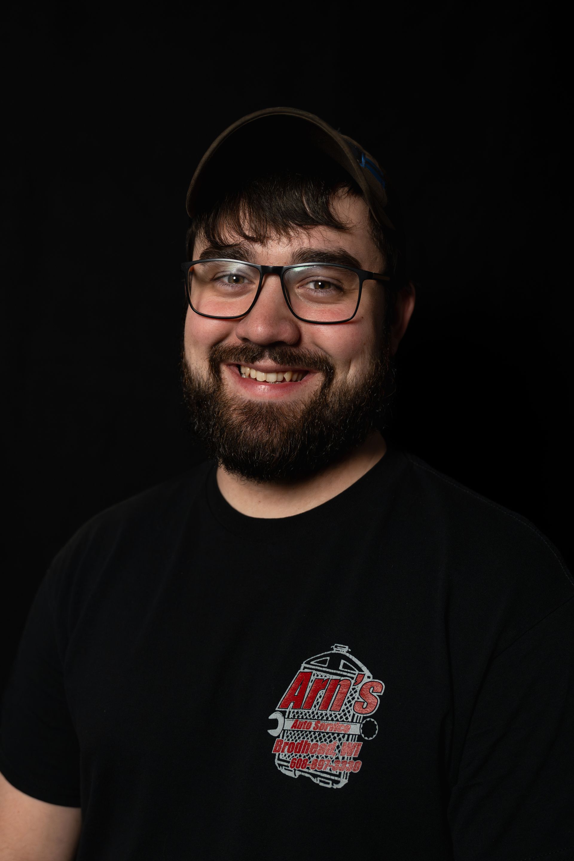 Man with glasses, shaved head, black shirt with logo, smiling against black background.  | Arn's Auto Service