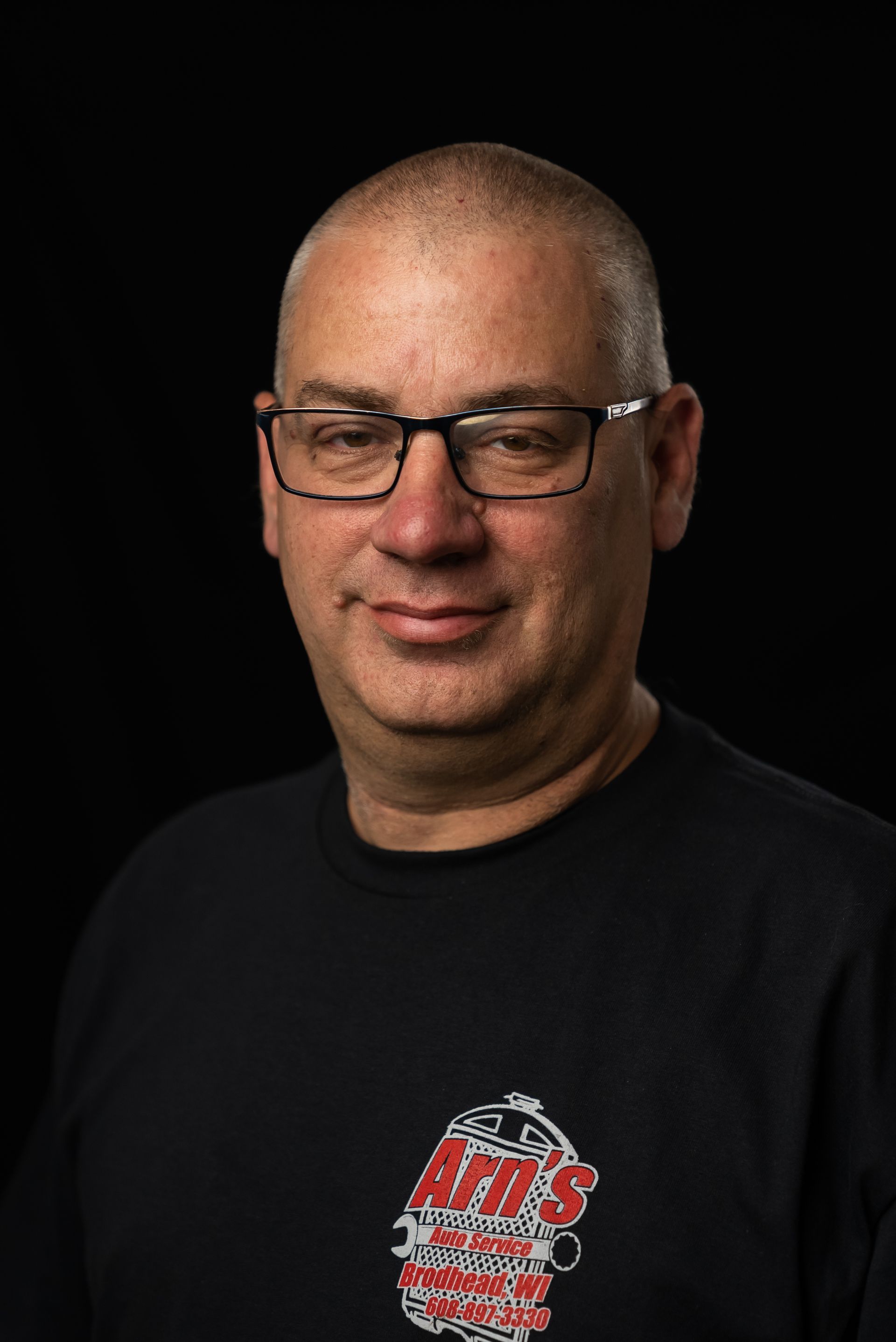 Man with glasses, shaved head, black shirt with logo, smiling against black background.  | Arn's Auto Service